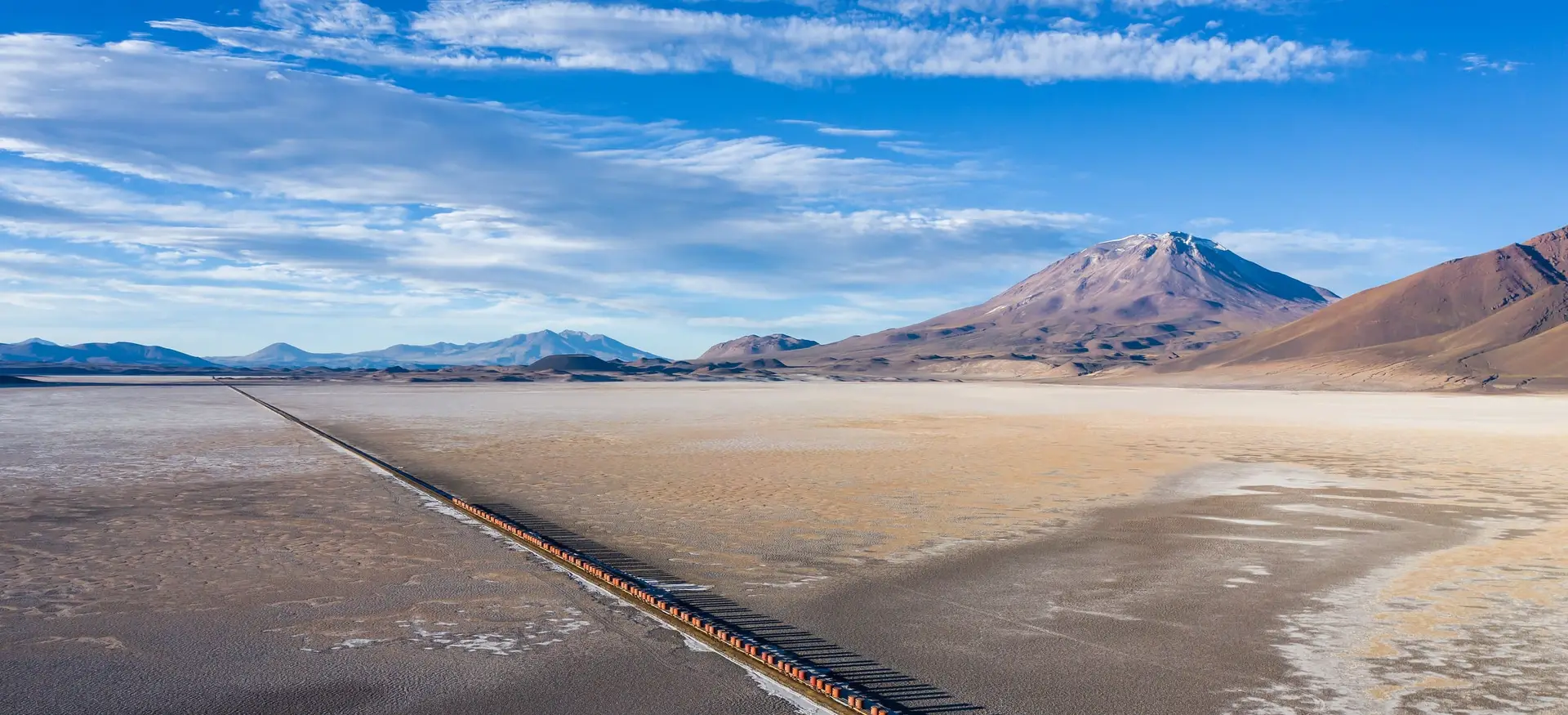 A long freight train crossing a vast salt flat towards a snow-capped volcano under a bright blue sky.