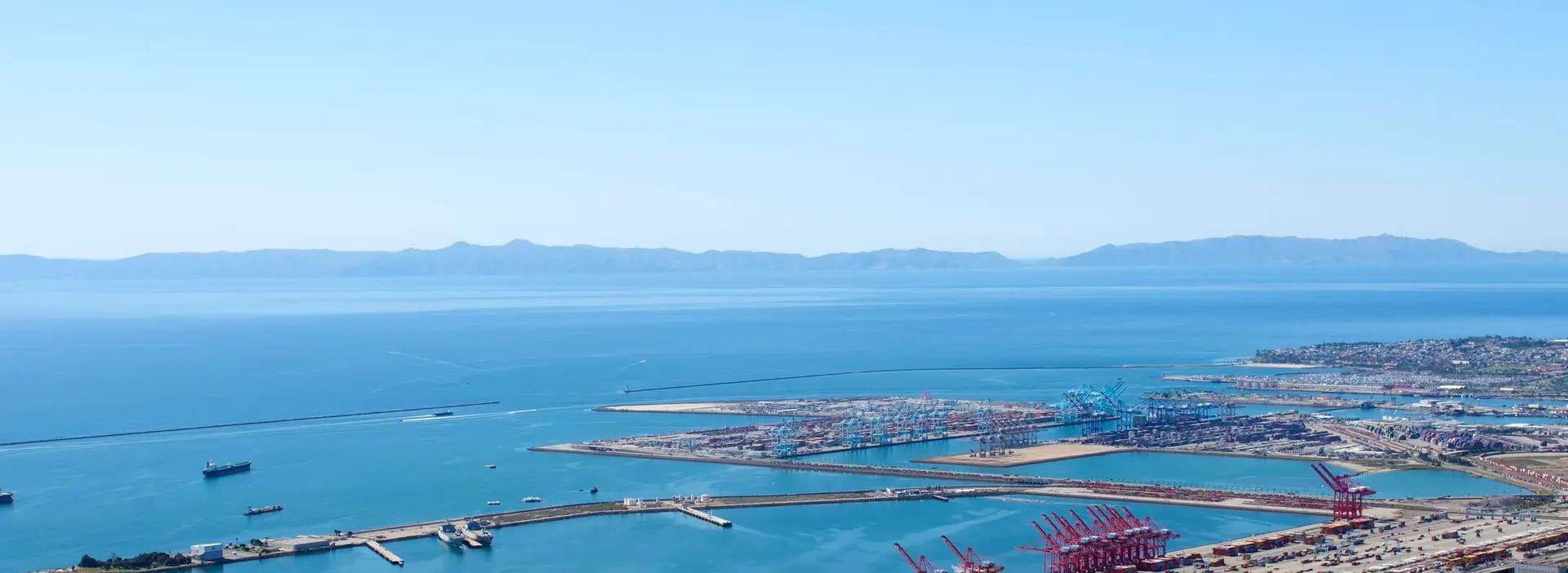 A wide view over a sunlit container port and harbour breakwaters, with islands on the horizon.
