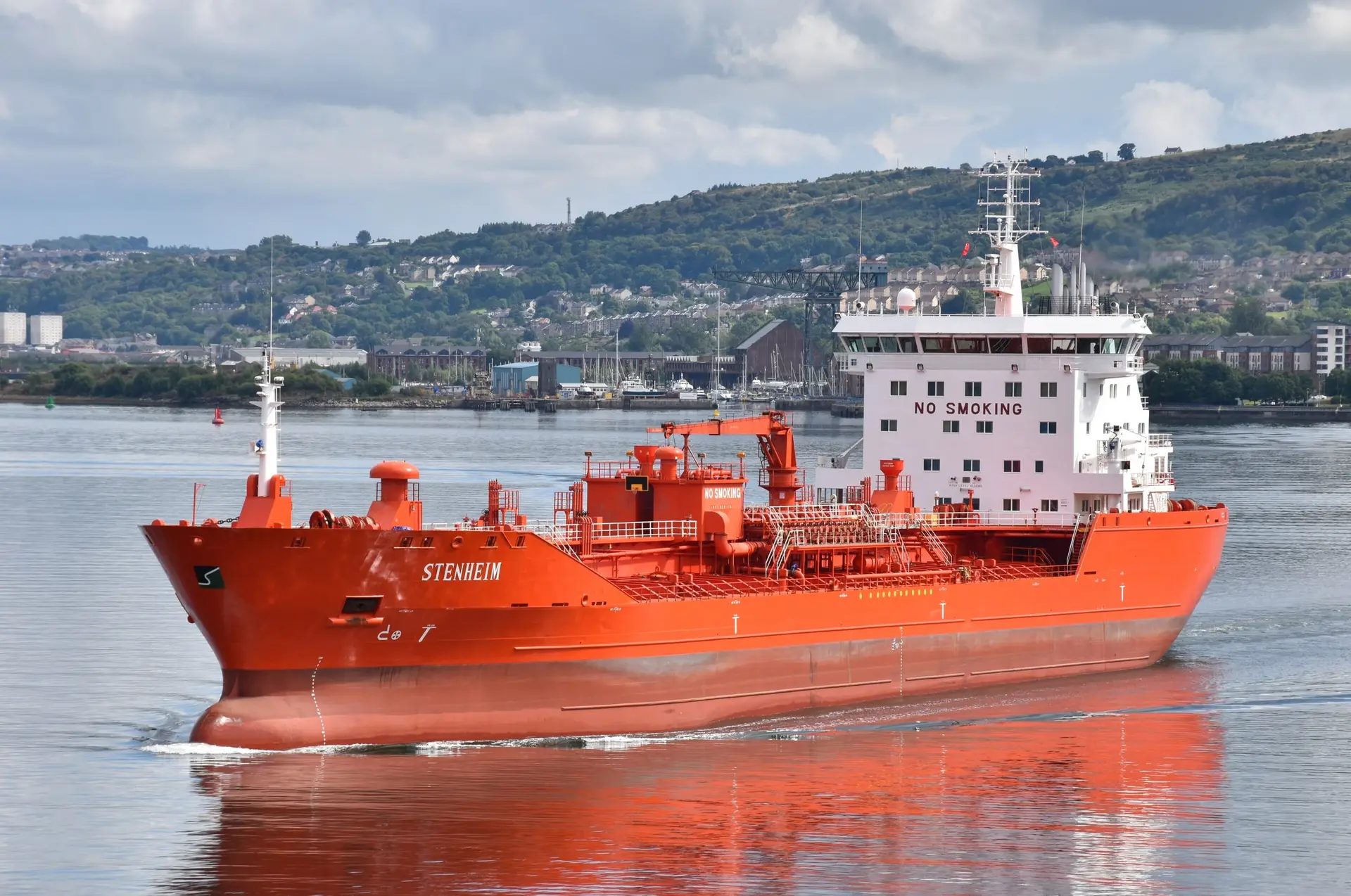 A bright orange chemical tanker under way on calm water, with green hills behind.