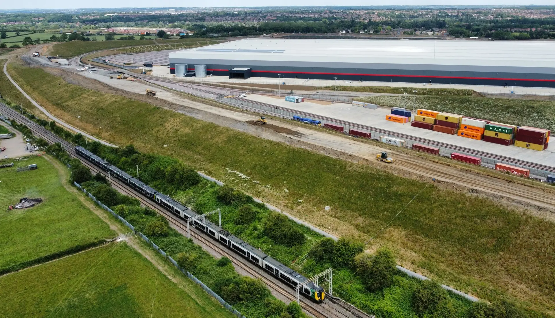 A large distribution warehouse beside a rail-served container terminal, seen from the air across green fields.
