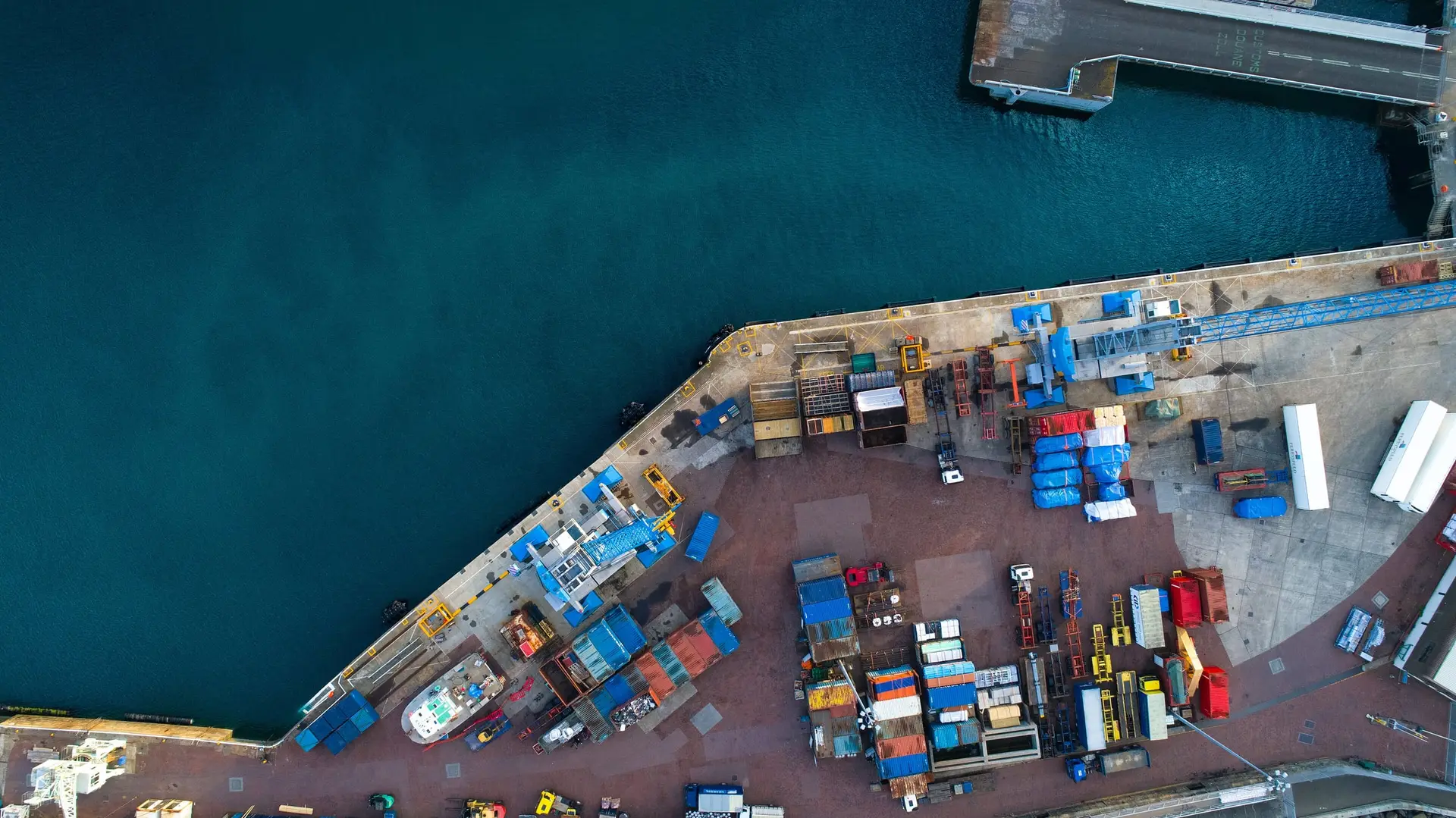 A harbour quay from directly above, with containers and cargo staged beside deep blue water.