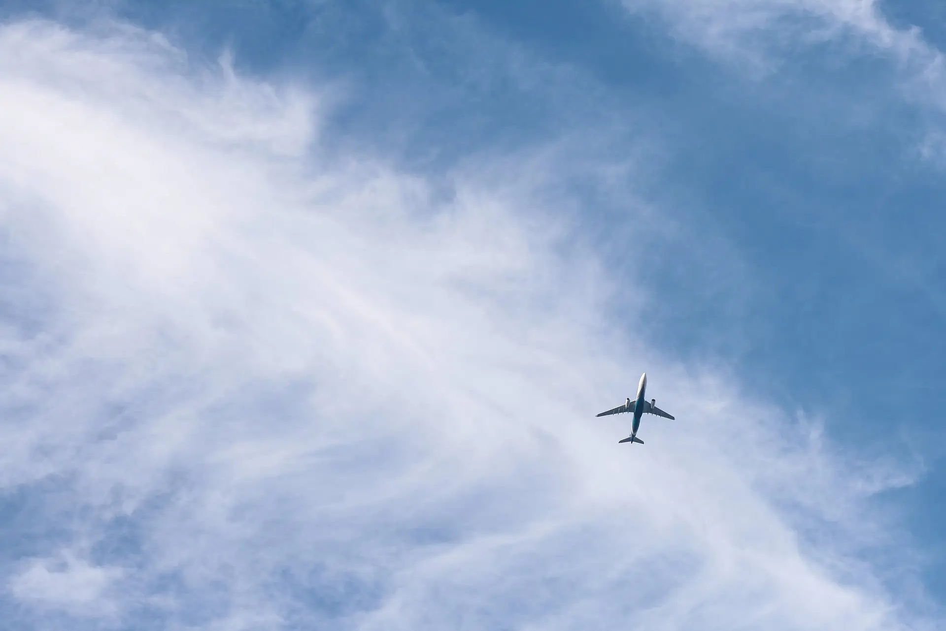 A jet aircraft high in a bright blue sky streaked with white cloud.